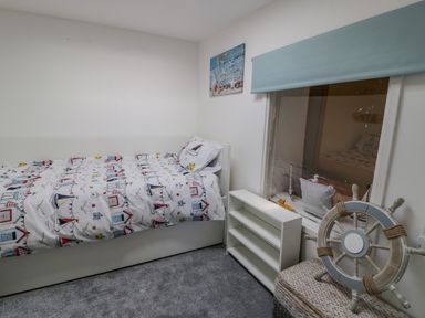 A small bedroom with a single bed with nautical themed bedding a white shelf under a window and a ship wheel decoration in the corner at Penrallt in Cemaes Bay