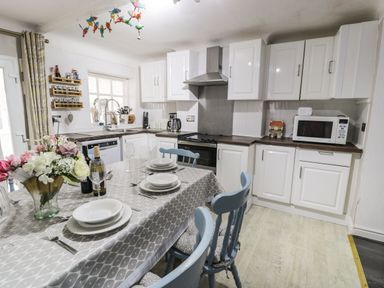 A kitchen with white cabinets a dining table set with plates and glasses and blue chairs at Penrallt in Cemaes Bay