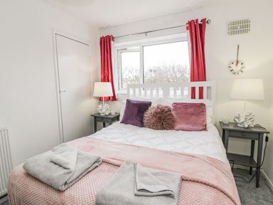 A bedroom with a white bed with pink and purple pillows two bedside tables with lamps and coral decorations and red curtains at Penrallt in Cemaes Bay