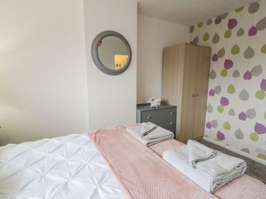 A bedroom with a bed that has folded towels on a pink throw a chest of drawers with a decorative item a wooden wardrobe and a circular mirror on the wall at Penrallt in Cemaes Bay