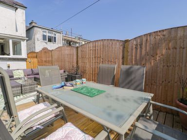 A wooden fenced outdoor patio with a glass table, chairs with cushions, and a wicker sofa set at Penrallt in Cemaes Bay