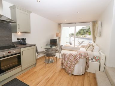 A kitchen and living area with a glass dining table chairs a white sofa and large window overlooking water at The Moorings in Porthmadog