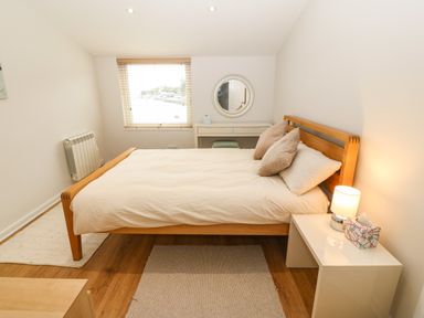 A bedroom with a wooden bed a white bedside table with a lamp and tissue box a dressing table with a round mirror and window blinds at The Moorings in Porthmadog
