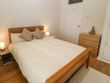 A bedroom with a wooden bed white bedding two pillows two bedside tables with lamps a wooden chest of drawers with a statue and a white door at The Moorings in Porthmadog