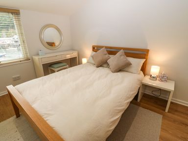A bedroom with a wooden bed, white bedding, two pillows, a white nightstand with a lamp and tissue box, a dresser with a round mirror and a stool at The Moorings in Porthmadog
