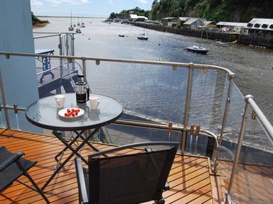 A balcony with a glass table holding two white cups a coffee press and a plate of cherry tomatoes overlooking a river with boats at The Moorings in Porthmadog