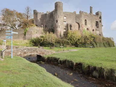 Stone bridge over a small stream with a castle ruin and green grass at Mews Cottage in Laugharne