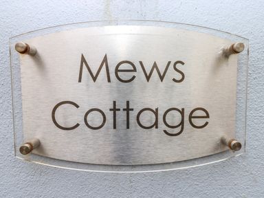 A metal and glass sign reading Mews Cottage mounted on a light blue textured wall at Mews Cottage in Laugharne