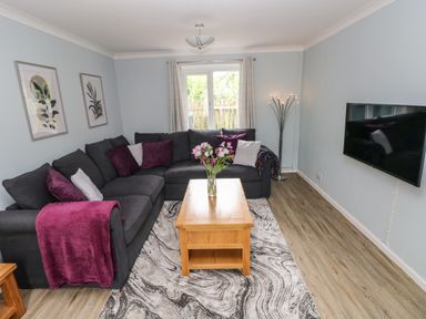 A living room with a gray sectional sofa, a wooden coffee table with flowers, a wall-mounted TV, and a floor lamp at Mews Cottage in Laugharne