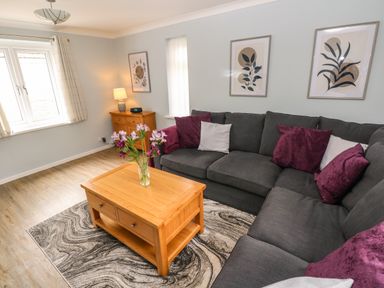 A living room with a dark grey sectional sofa with purple and white cushions a wooden coffee table with flowers a wooden cabinet with a lamp and framed leaf art on the walls at Mews Cottage in Laugharne