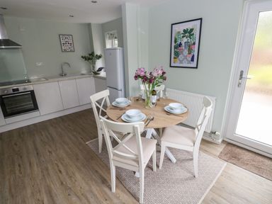 A kitchen and dining area with a round wooden table set for four with white chairs and flowers a white fridge and countertop with an oven and sink Mews Cottage in Laugharne