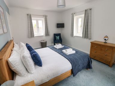 A bedroom with a bed with blue bedding two windows a wooden dresser and a blue chair at Mews Cottage in Laugharne