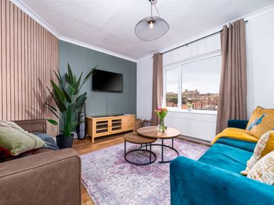 A living room with a blue sofa and brown armchair facing a TV on a wooden stand with a window showing a town view at The View in Whitby