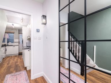 A hallway with a large grid mirror reflecting a staircase and a rug on wooden floor leading to a kitchen with white cabinets at The View in Whitby