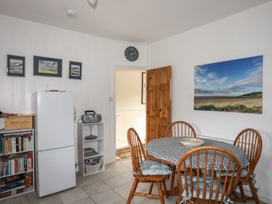 A dining area with a round table and four wooden chairs a fridge and a bookshelf in the corner at Amelie Cottage in Beaumaris Anglesey