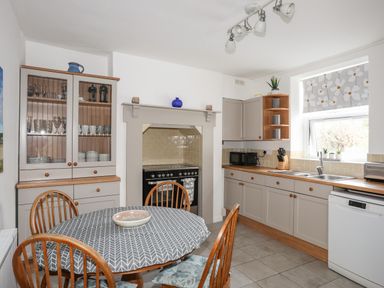 A kitchen with a round dining table covered by a patterned tablecloth with wooden chairs cupboards a stove a window with a floral blind and kitchen appliances at Amelie Cottage Beaumaris Anglesey