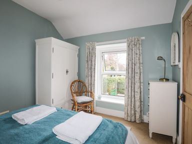 A bedroom with a bed covered by a teal blanket and white towels a white wardrobe a wicker chair a window with floral curtains and a white dresser with a lamp at Amelie Cottage Beaumaris Anglesey