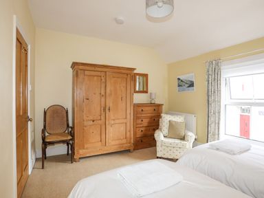 A bedroom with two single beds a wooden wardrobe a wooden chest of drawers a cushioned armchair and a window with floral curtains at Amelie Cottage in Beaumaris Anglesey