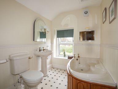A bathroom with a toilet a pedestal sink a corner bathtub and a window with a roller blind at Amelie Cottage in Beaumaris Anglesey