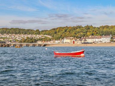 A red boat floating on water with a pier and houses on a hillside in the background at Amelie Cottage in Beaumaris Anglesey