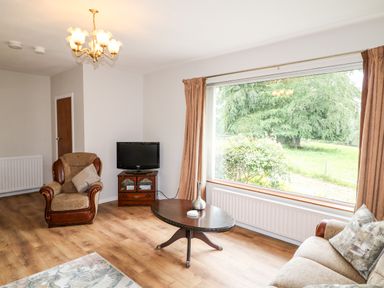 A living room with an armchair a sofa a coffee table a television and a large window showing green trees at Glebe Farm Cottage in Rathfriland County Down