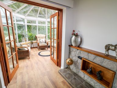 A conservatory with chairs and plants viewed through open wooden glass doors from a room with stone fireplace and decorative items at Glebe Farm Cottage in Rathfriland County Down