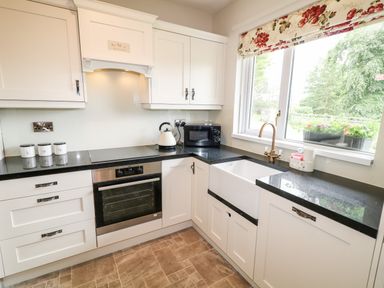 A kitchen with white cabinets black countertops an oven a kettle a microwave a farmhouse sink and a window with floral blind at Glebe Farm Cottage in Rathfriland County Down