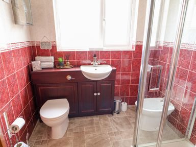A bathroom with a toilet, sink on a wooden cabinet, a glass shower enclosure, red tiled walls and a window at Glebe Farm Cottage in Rathfriland County Down