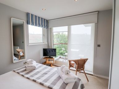 A bedroom with a bed, a mirror on the wall, a small TV on a stand, a wicker chair, and glass doors opening to a balcony at 9 Cedars in St Agnes