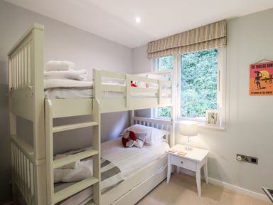 A bedroom with a white bunk bed next to a window and a small white bedside table with a lamp at 9 Cedars in St Agnes