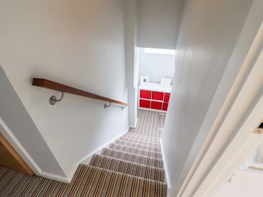 Carpeted stairs with a wooden handrail leading down to a landing with a white storage unit containing red fabric drawers and two framed pictures at 9 Cedars in St Agnes