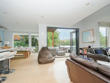 An open living room and kitchen area with brown leather sofas a bean bag dining table and chairs and a view to an outdoor patio with seating and hot tub at 9 Cedars in St Agnes