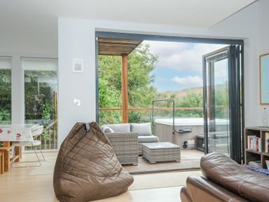 A seating area with a brown beanbag chair and leather sofa opening to an outdoor deck with wicker furniture and a hot tub at 9 Cedars in St Agnes