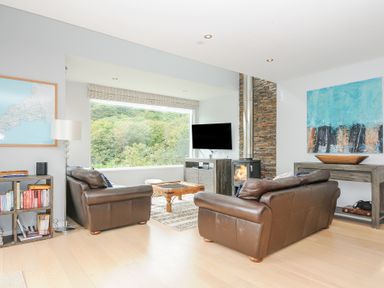 A living room with two brown leather sofas a wooden coffee table a wall-mounted TV and a window showing trees at 9 Cedars in St Agnes