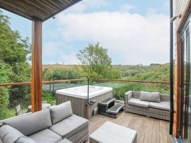An outdoor wooden deck with a hot tub and two grey cushioned sofas at 9 Cedars in St Agnes