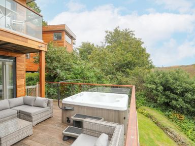 An outdoor wooden deck with a hot tub wicker sofas and table surrounded by trees at 9 Cedars in St Agnes