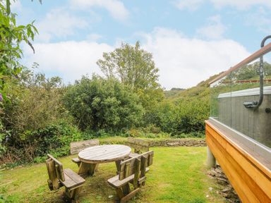 An outdoor seating area with a round wooden table and four wooden benches on grass near a wooden deck with glass railing at 9 Cedars in St Agnes