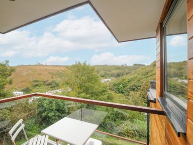 A balcony with a white table and chair overlooking green hills and trees at 9 Cedars in St Agnes