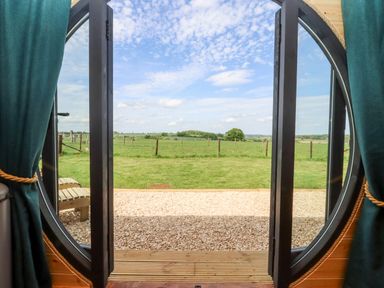 A round door opening to a gravel path and a grassy field with a fence and trees visible at The Happy Valley Pod in Chipping Norton