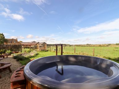An outdoor hot tub with benches and a garden swing in a grassy yard overlooking fields at The Happy Valley Pod in Chipping Norton