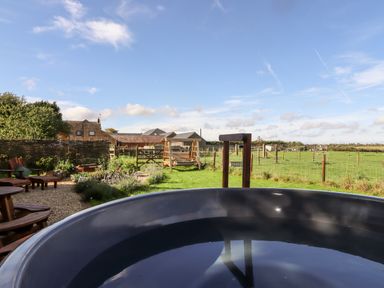 An outdoor garden with a large black tub in the foreground wooden chairs and tables on gravel a wooden swing and fenced green fields at The Happy Valley Pod in Chipping Norton