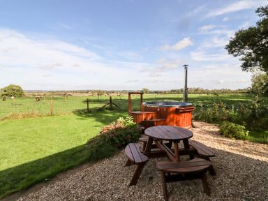 An outdoor area with a wooden picnic table and a hot tub overlooking a green field with cows at The Happy Valley Pod in Chipping Norton