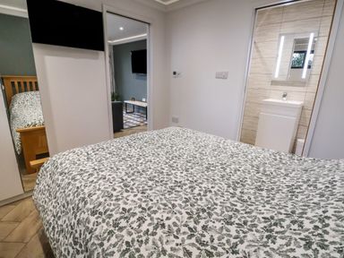 A bedroom with a bed covered in a leafy patterned bedspread a wall mounted tv mirrored wardrobes a glimpse into an adjoining room and a bathroom with changing wall tiles at The Nest in Prestatyn
