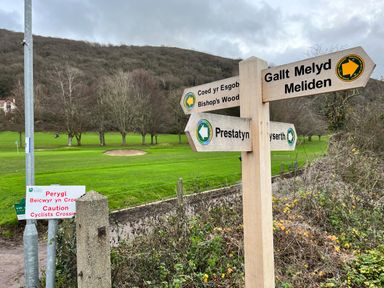 A wooden signpost showing directions to Bishop's Wood Meliden and Prestatyn near a green field with trees and a caution sign about cyclists crossing