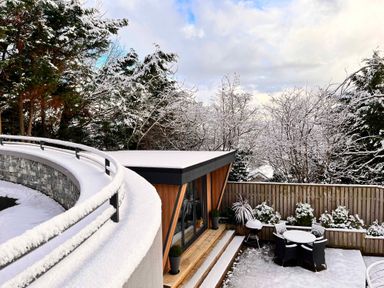 A snow-covered backyard with a small wooden structure a round table with chairs and trees in the background at The Nest in Prestatyn