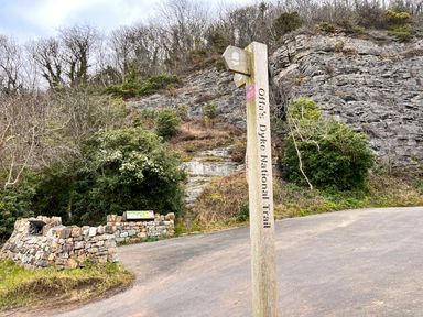 A wooden signpost reading Offas Dyke National Trail near a stone wall and rocky hillside with trees