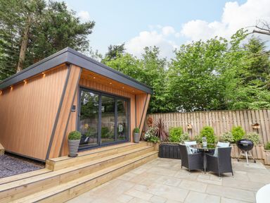 A wooden cabin with glass doors next to a patio with table and chairs and a barbecue at The Nest in Prestatyn