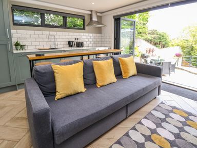 A kitchen area with green cabinets and white tiled backsplash with a gray sofa and yellow cushions in front at The Nest in Prestatyn
