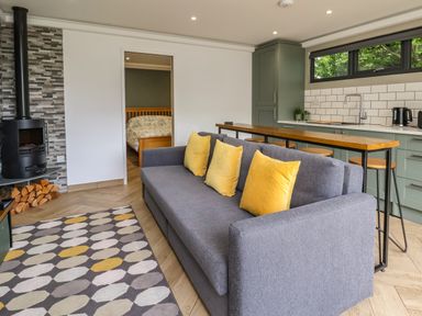 A living room with a gray sofa with yellow pillows and a kitchen area with a wooden countertop and bar stools at The Nest in Prestatyn