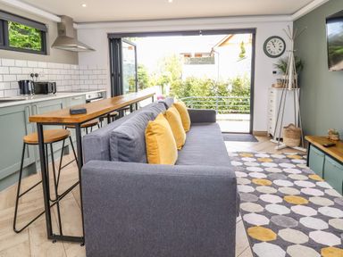 A living room with a gray sofa with yellow pillows a wooden bar table with stools kitchen appliances a patterned rug a wall clock and sliding glass doors at The Nest in Prestatyn